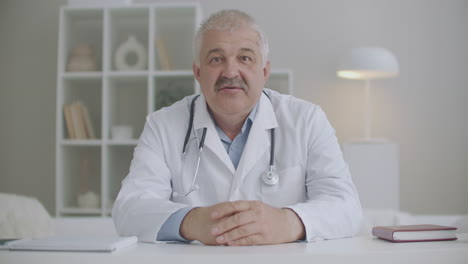 middle-aged-male-doctor-is-greeting-participants-of-webinar-or-video-conference-and-speaking-portrait-in-office-of-clinic